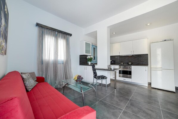 A modern living room with a red sofa, a glass coffee table, and a kitchen area with white cabinets and a refrigerator.