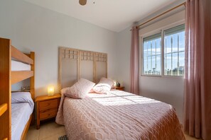 Un dormitorio con literas, una mesita de noche con lámpara, cabecera y ventana con cortinas.