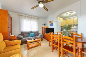 Una sala de estar con un sofá, una mesa de centro, una mesa de comedor con sillas, un ventilador de techo y una cocina visible a través de un arco.