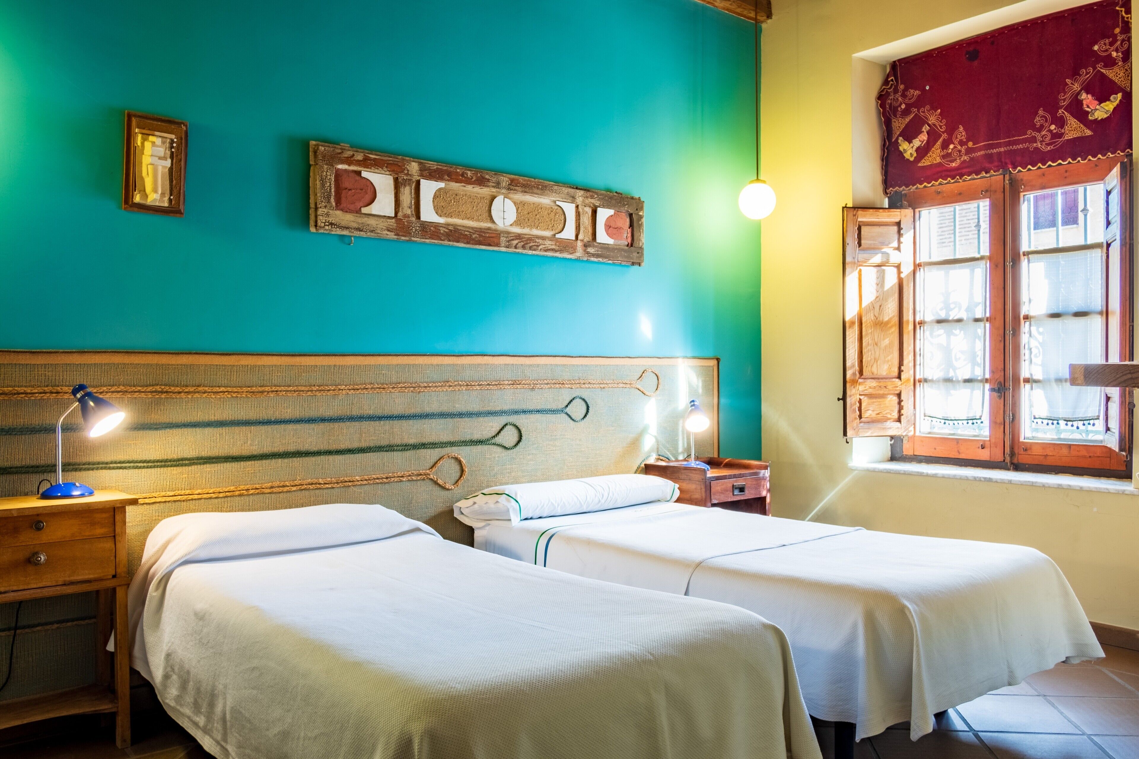 A room with two beds, a wooden headboard, a window with curtains, and a wall-mounted picture frame.