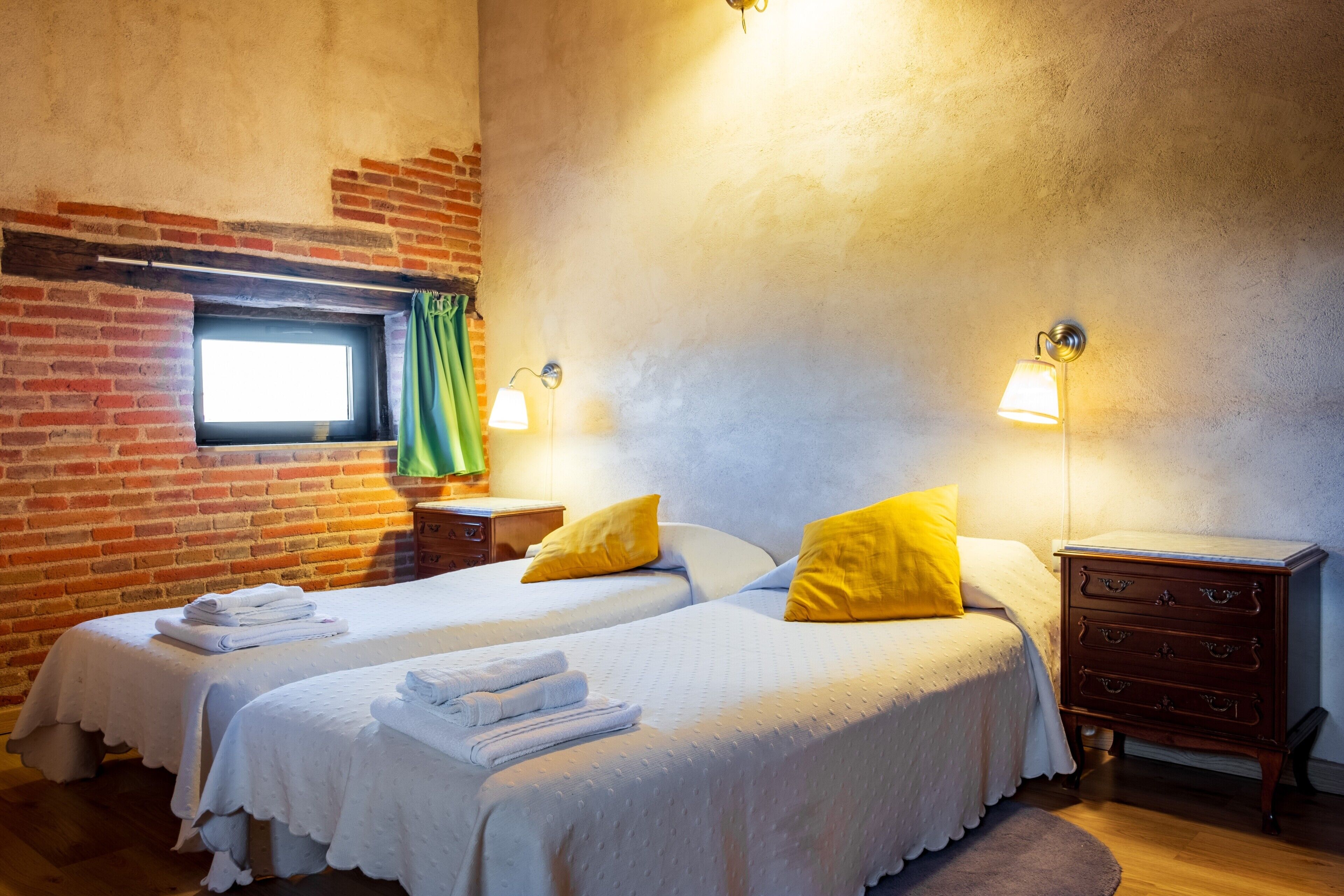 Une chambre avec deux lits, un mur en briques, une fenêtre avec des rideaux et une commode en bois.
