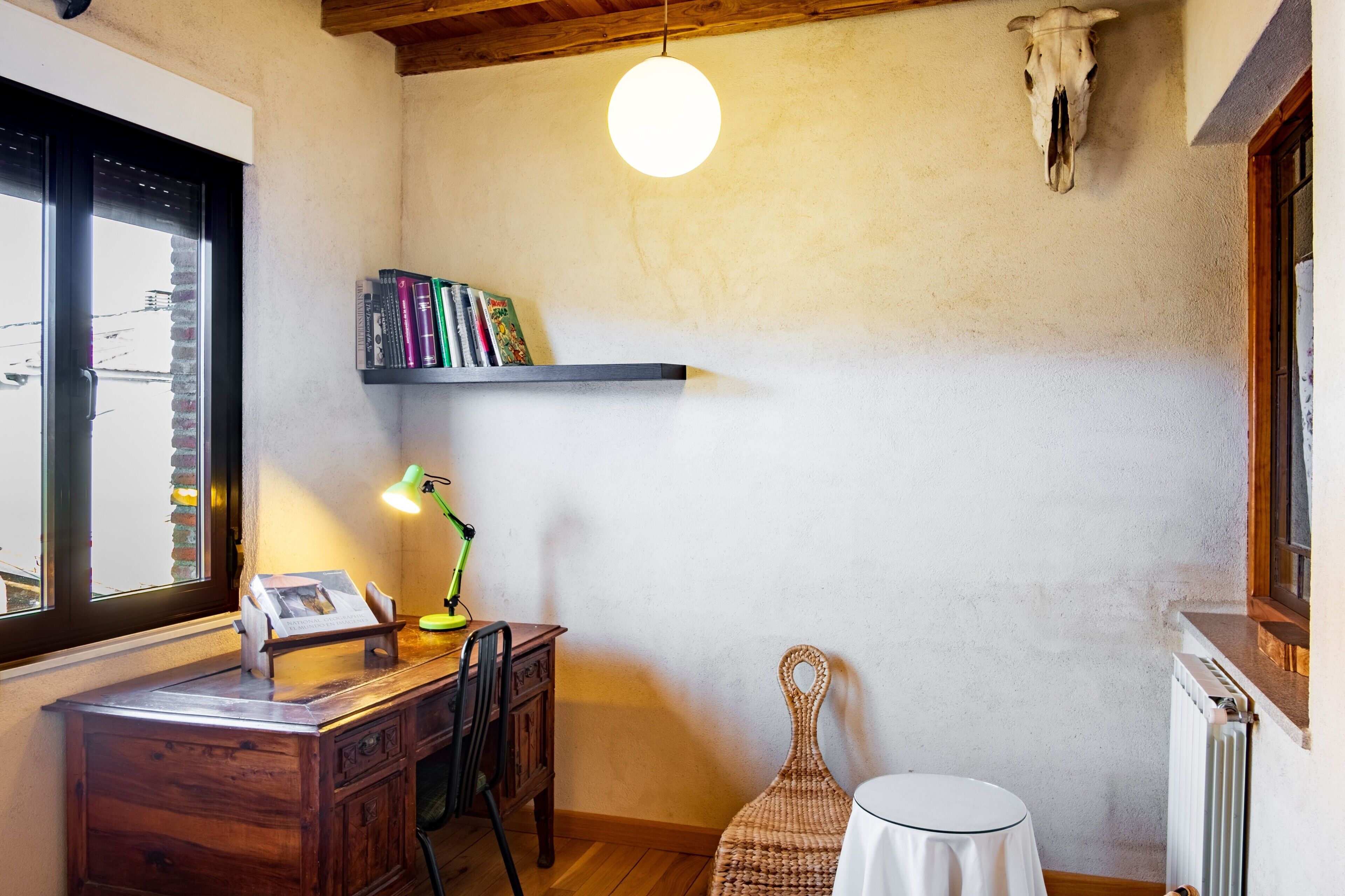 Una stanza con una scrivania in legno, una sedia, un tavolino, un lampadario, uno scaffale con libri e un teschio appeso al muro.