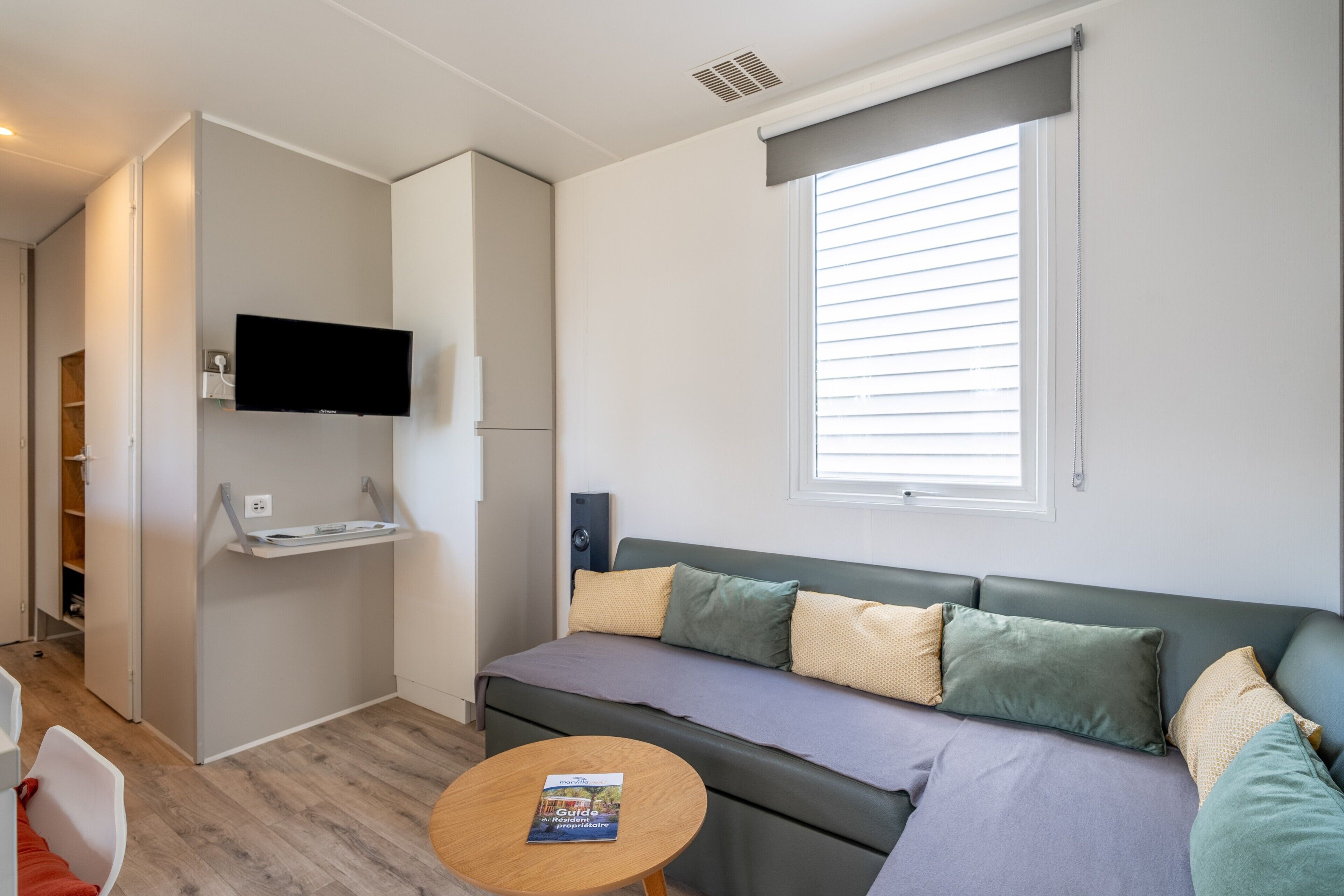 Un espacio habitable funcional con sofá, una mesita, televisor montado en la pared y ventana con persianas.
