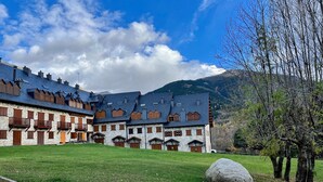 A traditional stone building with multiple balconies and a stone facade, set against a backdrop of mountains and a clear sky.