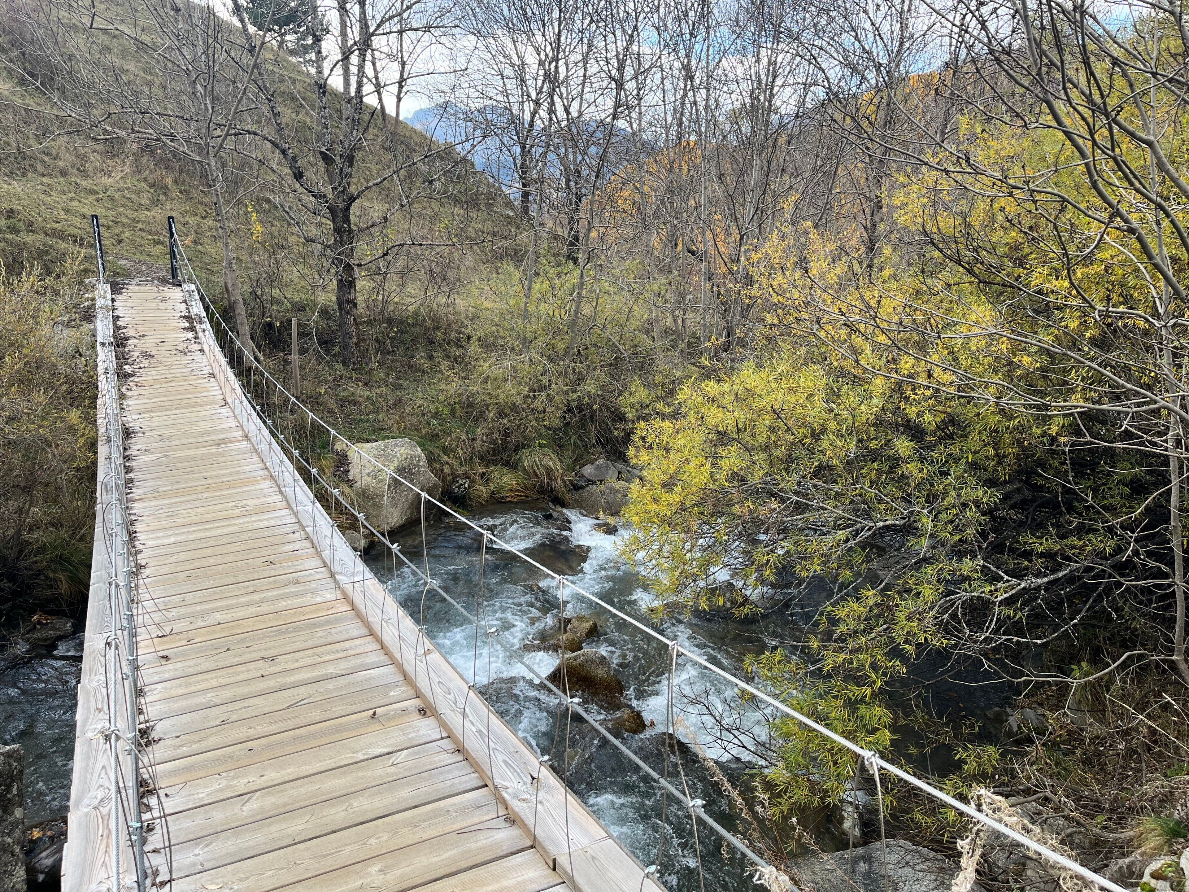 Uma ponte suspensa de madeira sobre um rio corrente, cercada por árvores e terreno rochoso.