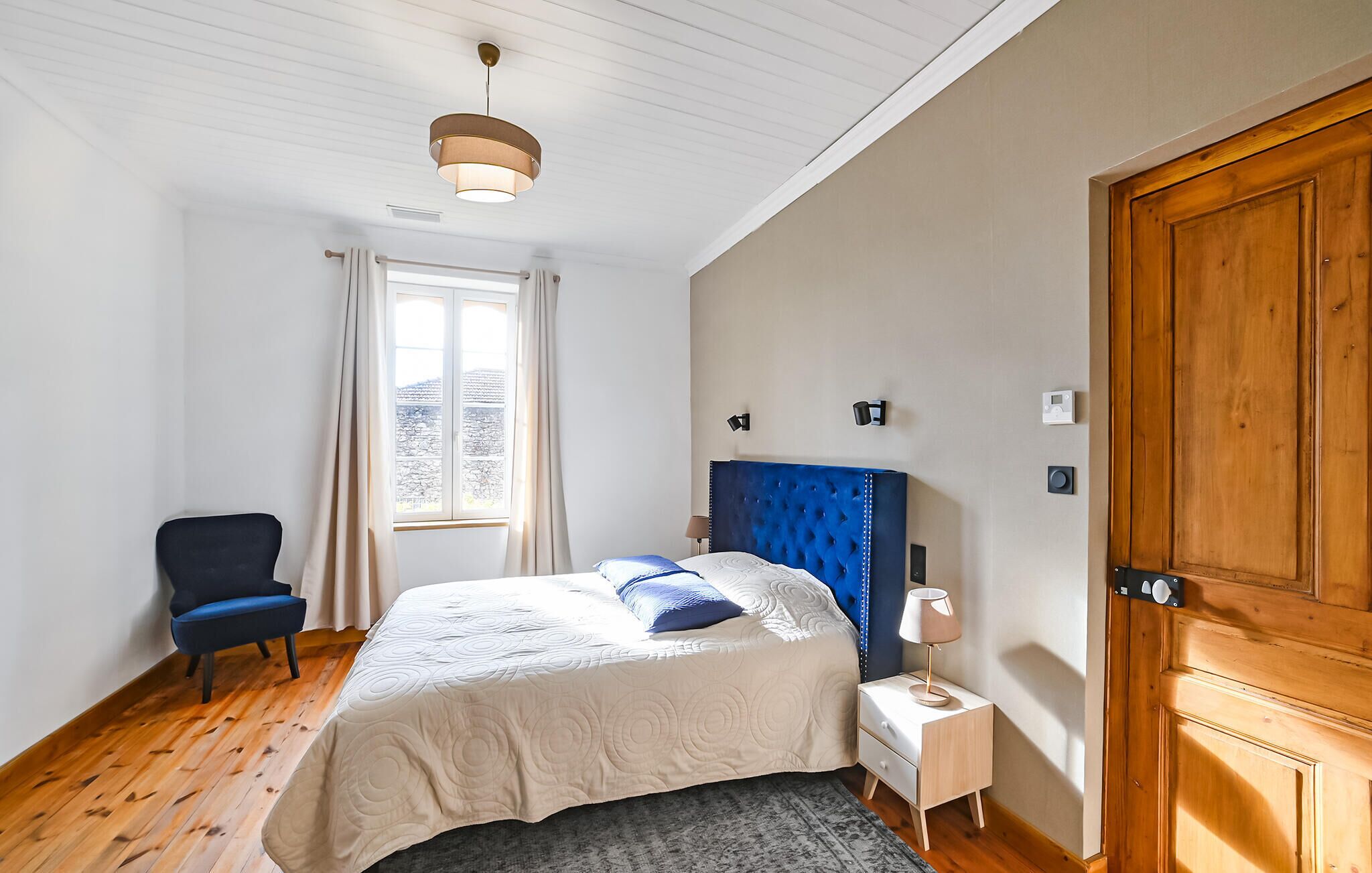 Un dormitorio con cama, una silla, una mesita de noche, una lámpara, una puerta de madera y una ventana con cortinas.