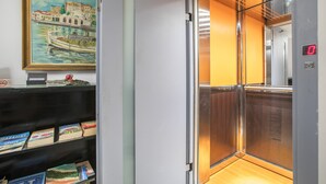 An elevator with a digital display, a painting of a canal scene, and a shelf with books.