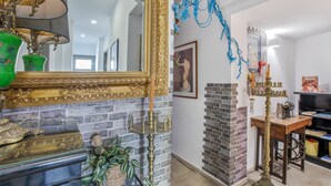 Una habitación con pared de ladrillo, un espejo grande y ornamentado, un jarrón verde, un candelabro de latón y un cuadro enmarcado en la pared.