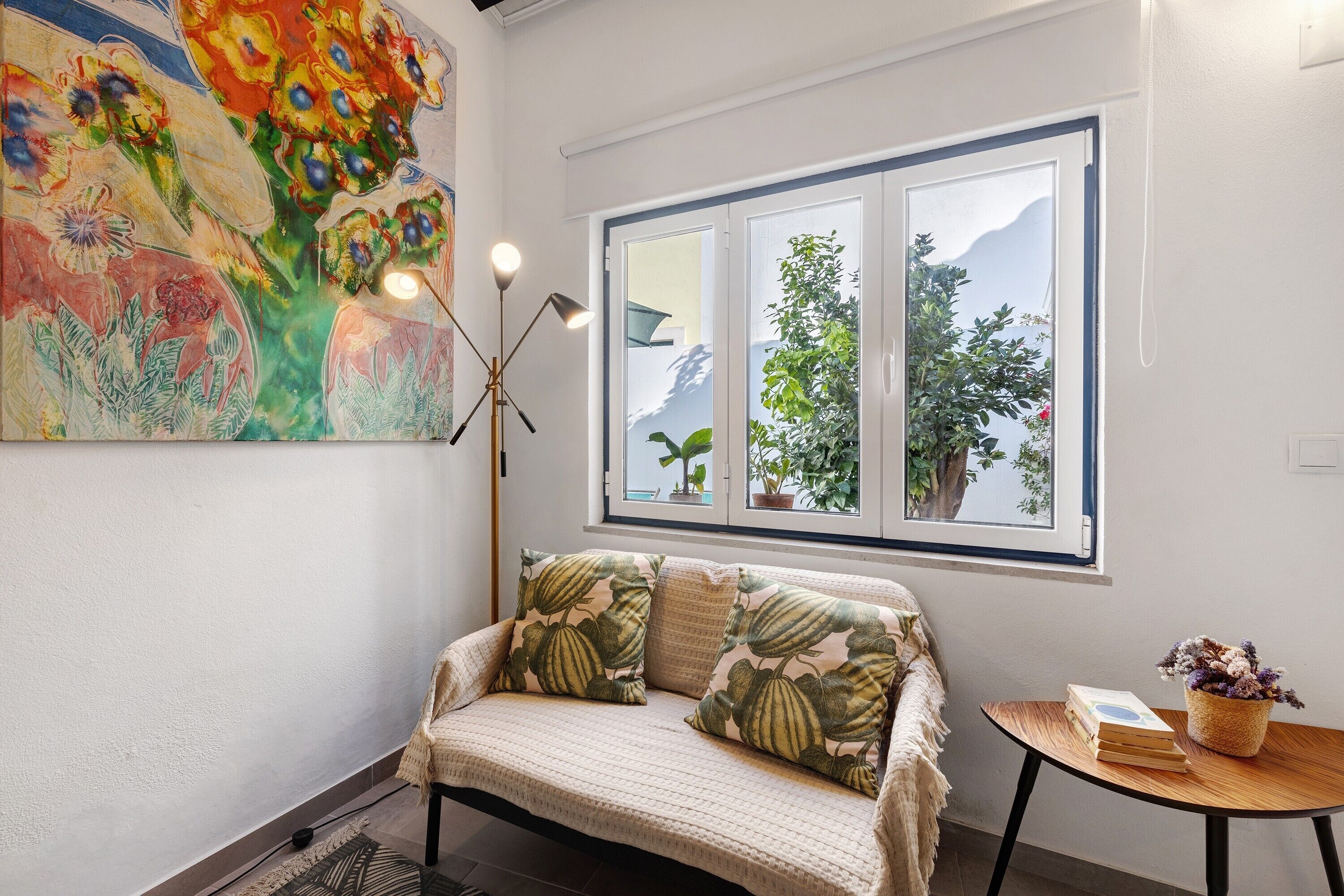 Una habitación acogedora con sofá, ventana con vistas a la naturaleza, un cuadro en la pared y una mesita con un libro y un jarrón.
