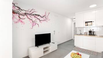 A modern kitchen with white cabinetry, a flat-screen TV, and a fruit bowl on a white table.