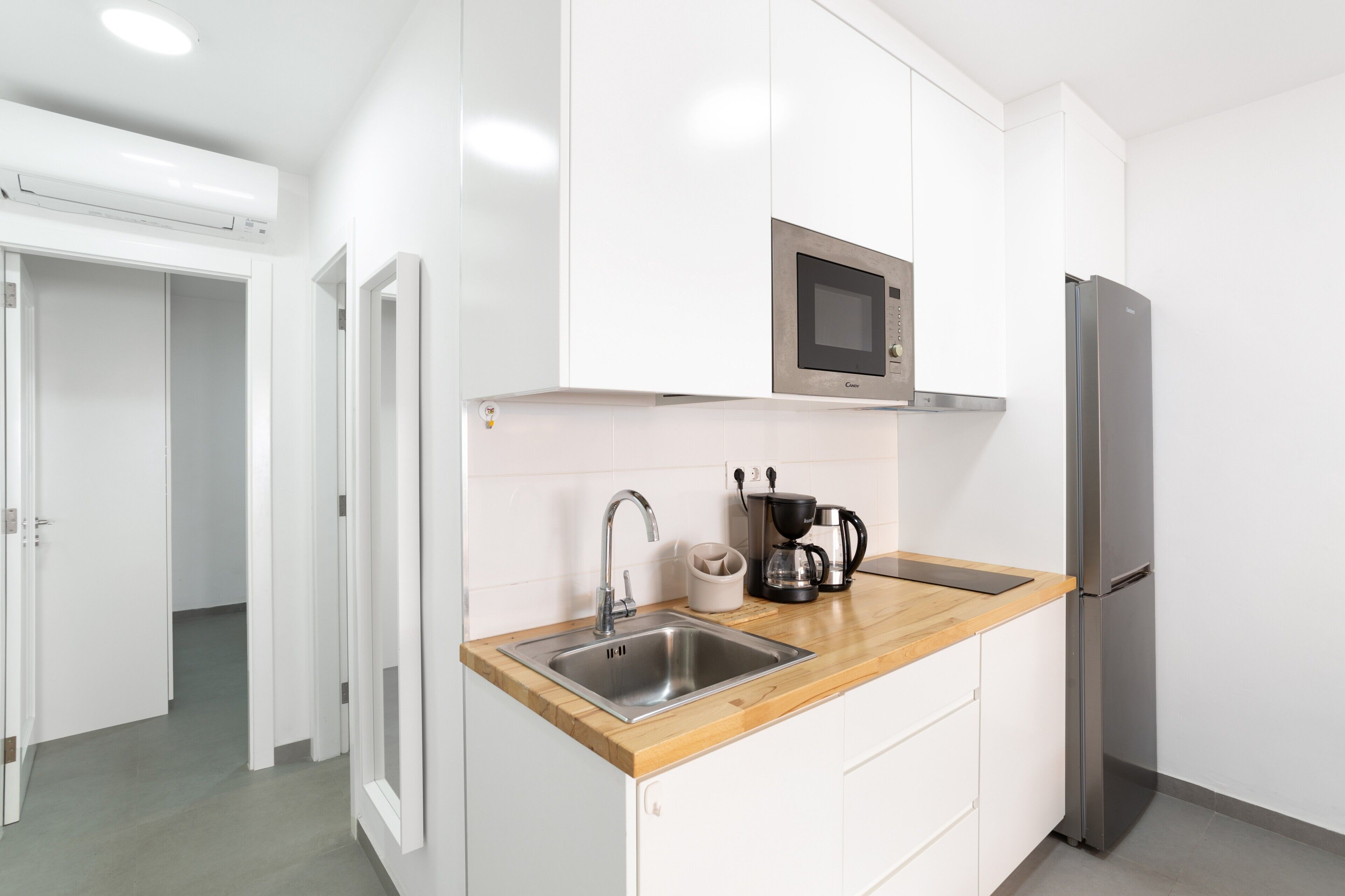 Una cocina moderna con armarios blancos, un lavamanos de acero inoxidable, un microondas, una cafetera y un refrigerador.