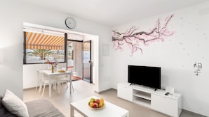 Una sala de estar moderna con un sofá, una mesa de comedor con sillas, un televisor sobre un mueble bajo y una pared con un mural de cerezos.