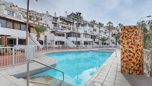 Una piscina con solárium empedrado, rodeada de edificios blancos con balcones y palmeras.