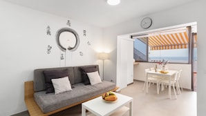 Una sala de estar moderna con un sofá, una mesa de comedor con sillas, una pequeña mesa de centro, un reloj de pared y un ventanal con toldo a rayas anaranjadas.