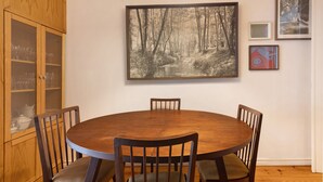 A dining area with a round wooden table, four chairs, a wooden cabinet with glass doors, and a framed forest painting on the wall.