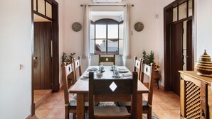 A dining area with a wooden table set for six, flanked by wooden chairs, a window with curtains, and a wall-mounted air conditioner. - Holiday Home 'Solar Da Tirosa' with Private Pool, Private Terrace and Wi-Fi (Boliqueime)