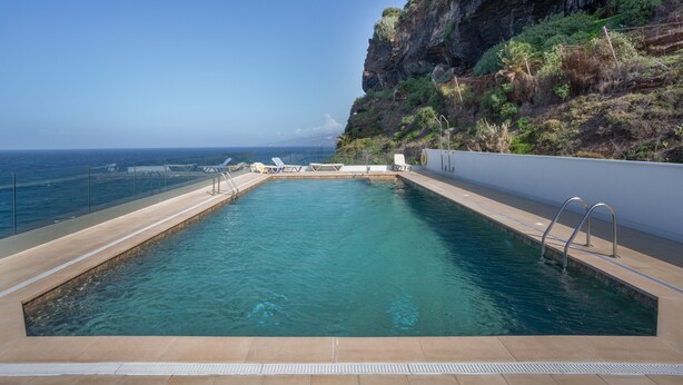 Una piscina rectangular amb aigua cristal·lina, envoltada per una paret blanca i un penya-segat rocos.