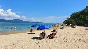 A beach with clear skies, people swimming and relaxing under umbrellas, and a chair with a striped towel. - Villa Daniella - Beach Homes (Florianópolis)