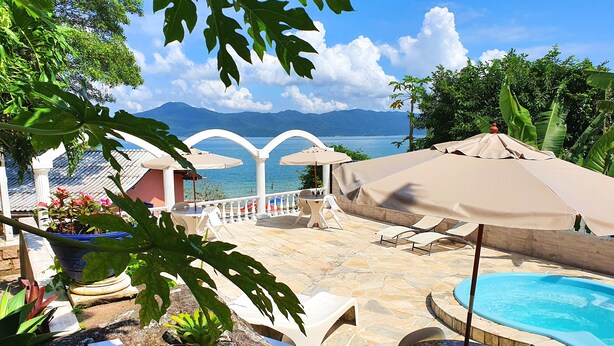 A pool area with lounge chairs and umbrellas, a view of the sea, and a mountainous backdrop. - Villa Daniella - Beach Homes (Florianópolis)