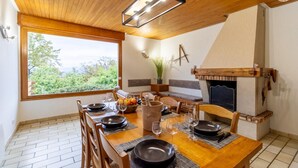 Un coin repas avec une table en bois dressée pour six personnes, une cheminée et une grande fenêtre donnant sur un paysage verdoyant.