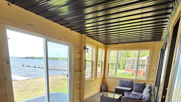 A wooden cabin with a large sliding glass door leading to a deck overlooking a lake. Inside, there is a gray sofa and wicker furniture. - Home on Grand Lake, in Saginaw, Near Ivy Black (Saginaw)