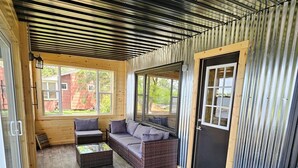 A covered patio area with a sofa, a small table, and a wicker chair. There are windows providing natural light and a door with a metal frame. - Home on Grand Lake, in Saginaw, Near Ivy Black (Saginaw)
