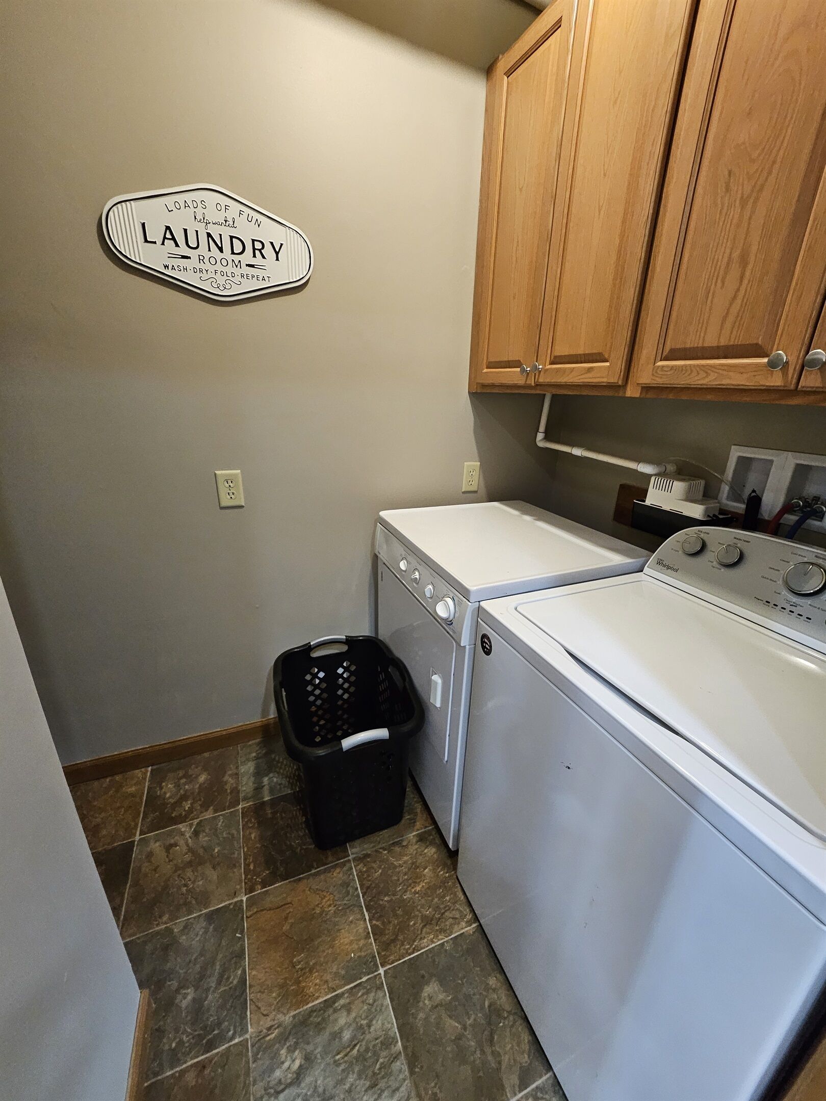 A laundry room with a sign indicating it's a laundry room, a washing machine, a dryer, wooden cabinets, and a tiled floor.