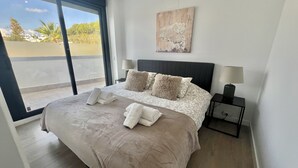 Una cama bien hecha con un amplio ventanal, una mesita de noche con lámpara y un cuadro en la pared.
