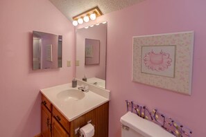 Un baño con una pared rosa, un lavabo blanco, armarios de madera, un espejo y un inodoro.