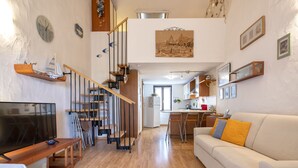 A modern living room with a wooden staircase, a flat-screen TV, a sofa with yellow cushions, and a kitchen area in the background. - Le Conchiglie by Interhome (Olbia)