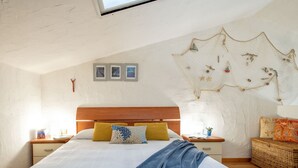 A bedroom with a large bed, wooden headboard, and a ceiling with a skylight. There are three framed pictures on the wall, a decorative wall hanging, and a wicker basket on the floor. - Le Conchiglie by Interhome (Olbia)
