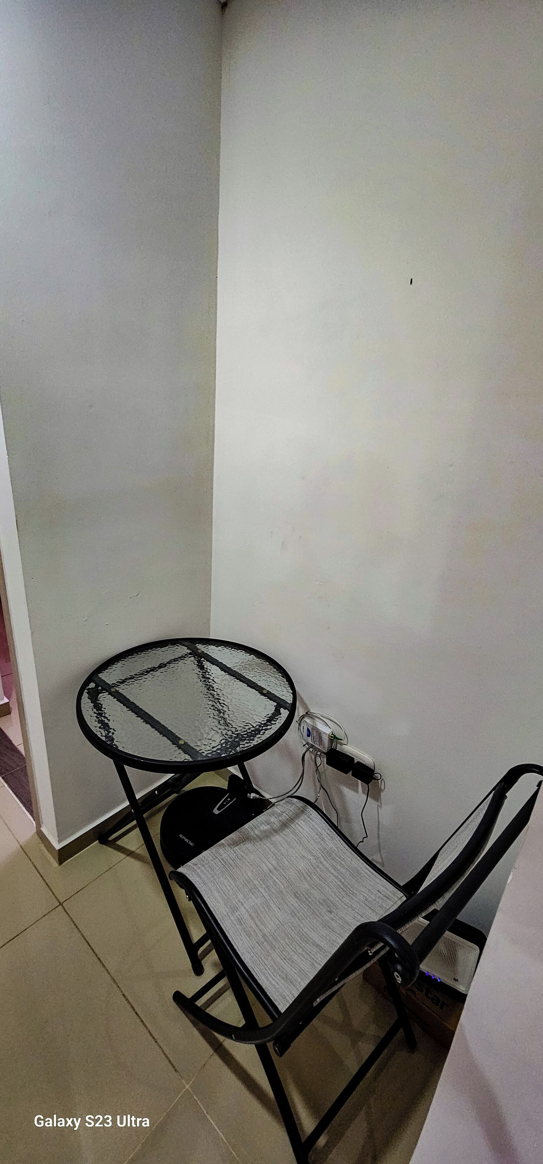 A corner room with a glass-top table and a chair, a power outlet on the wall, and a Galaxy S23 Ultra phone on the floor.