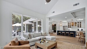 A spacious living room with a large window, a sofa, a coffee table, and a kitchen area with bar stools and a dining table. - Spacious Coastal Getaway | Private Pool, Lagoon Views with Dock, Balcony, and Yard (Hilton Head Island)