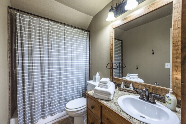 Un baño con cortina de ducha a rayas, un inodoro blanco, un tocador de madera con lavamanos y un espejo grande con luces arriba.