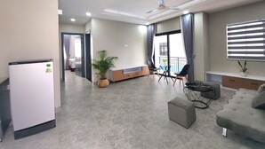 A modern living room with a sofa, ottoman, and a dining table with chairs. There is a refrigerator, a potted plant, and a ceiling fan. - My Natural Villas by You Long (Siem Reap)