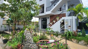 A modern multi-story building with a central staircase, surrounded by a landscaped garden with various plants and flowers. - My Natural Villas by You Long (Siem Reap)