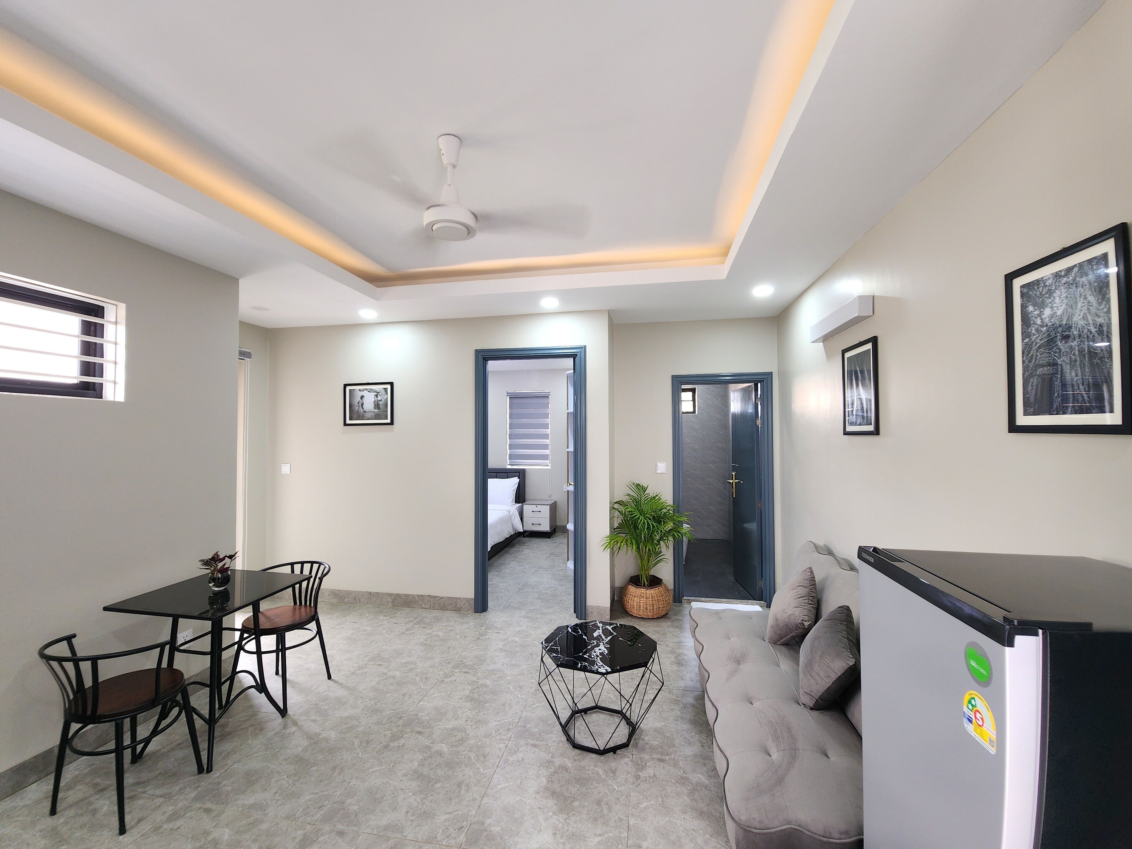 A modern living room with a sofa, a dining table with chairs, a coffee table, and a refrigerator. There is a door leading to another room and a window with blinds.