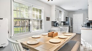 Una cocina moderna con armarios blancos, una mesa de comedor para cuatro personas y un ventanal con persianas.