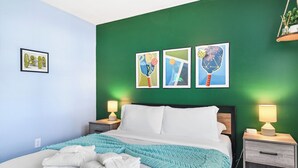 Una cama bien hecha con sábanas blancas y una manta turquesa, flanqueada por dos mesitas de noche con lámparas, en una habitación con paredes verdes y obras de arte enmarcadas.