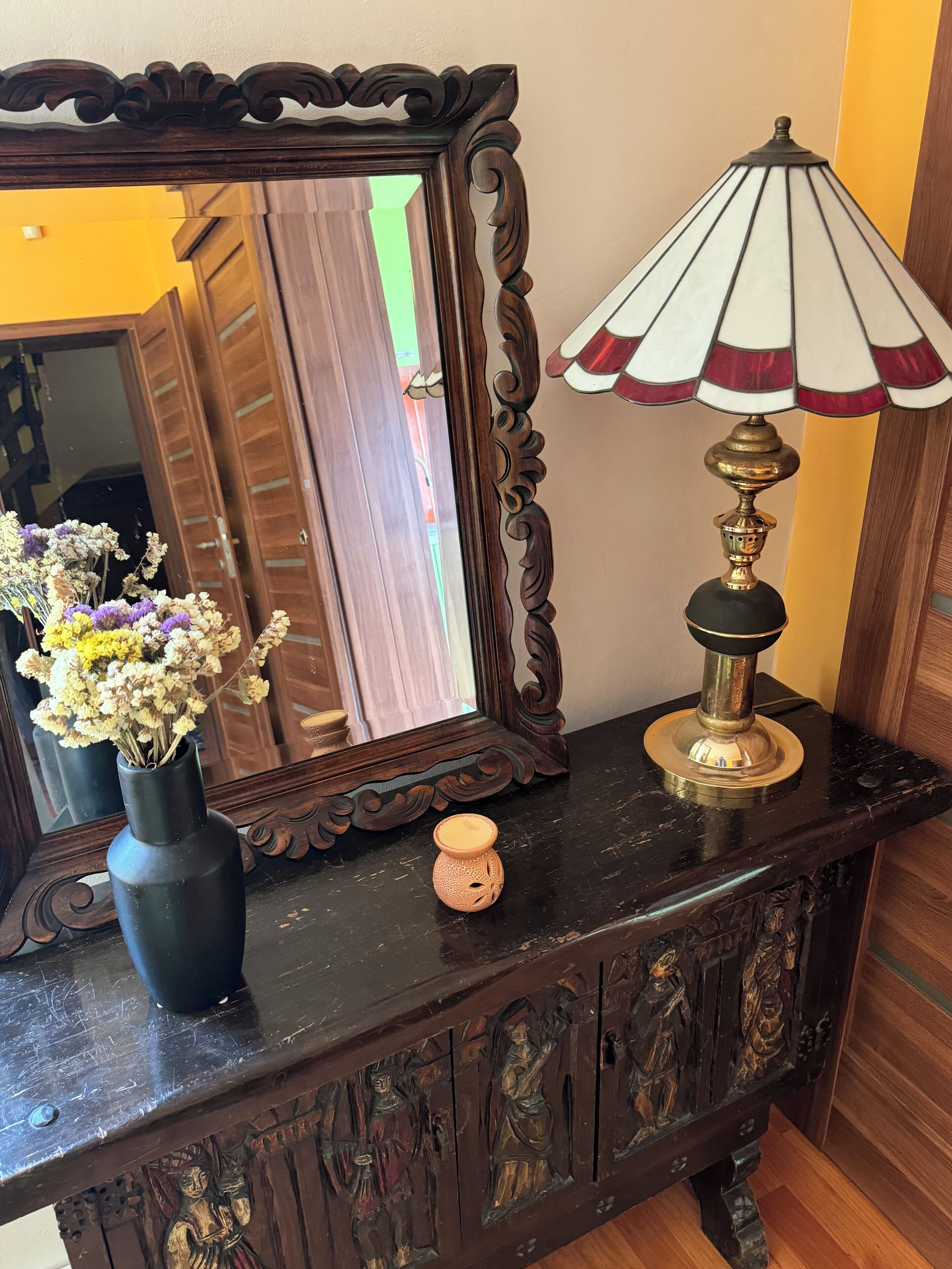 A wooden dresser with a mirror, a vase with dried flowers, and a table lamp.