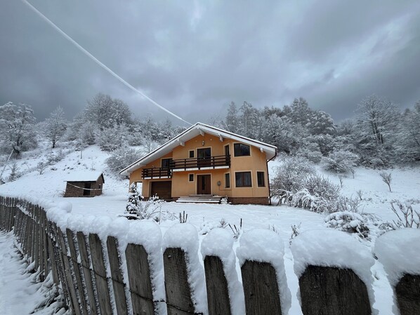 A two-story house with a balcony, surrounded by snow-covered trees and a wooden fence.