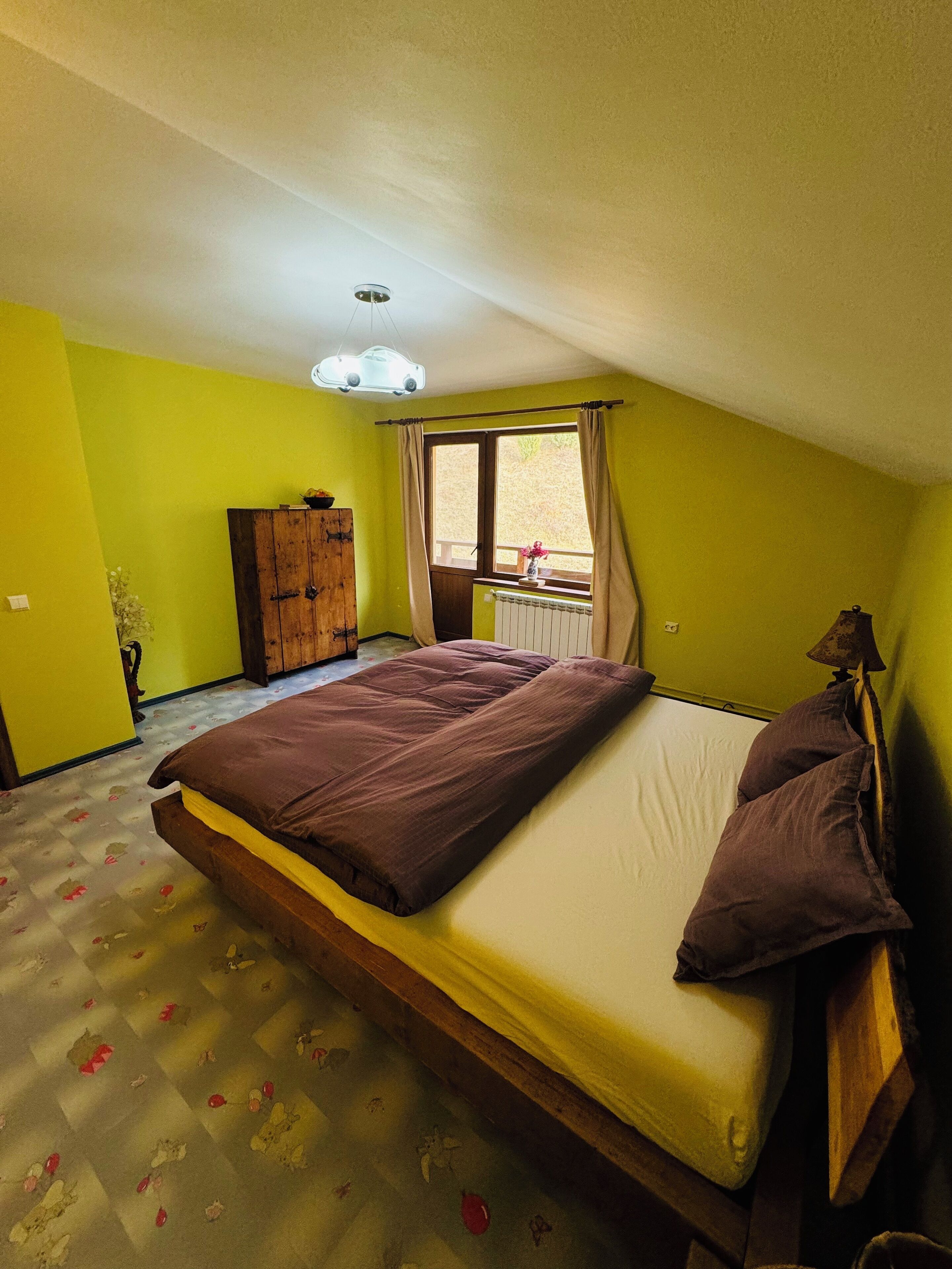 A bedroom with a bed, a window with curtains, a radiator, and a wooden wardrobe.