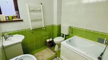 Une salle de bain avec un lavabo, des toilettes et une baignoire, des murs carrelés de vert et de blanc, et une fenêtre avec des stores.