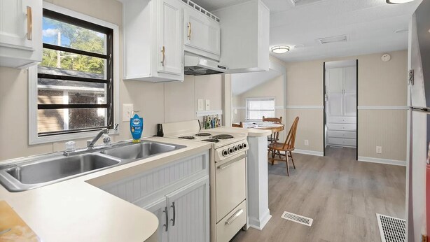 A modern kitchen with white cabinetry, a double sink, a stove, and a dining area with a table and chairs. - 1309 Ocean Lakes Pet Friendly Golf Cart⛱️🏝️🩱🌊👙☀️🐚 (Myrtle Beach)