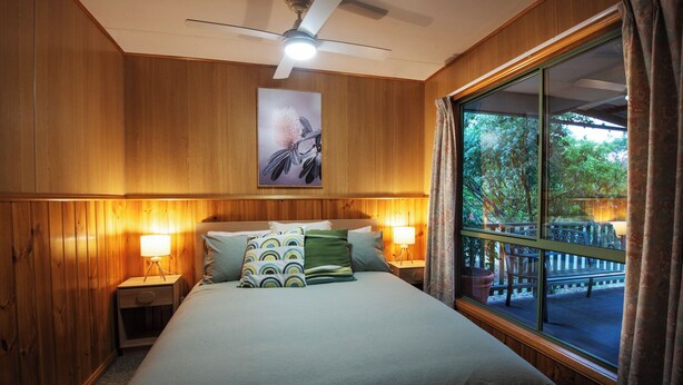 Een slaapkamer met houten lambrisering, een bed met geborduurde kussens, twee bedlampen, een plafondventilator en een groot raam met uitzicht op een terras.