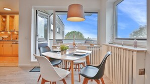 A modern dining area with a table set for two, a hanging light fixture, and large windows offering a view of the sky and buildings.