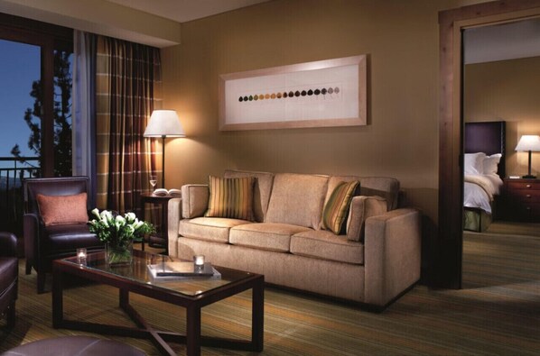 A hotel room with a sofa, armchair, and a coffee table. There is a framed artwork on the wall and a window with curtains.