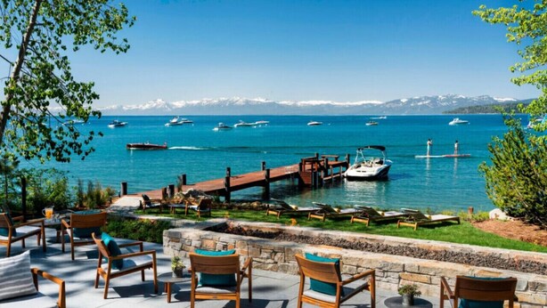 Une terrasse au bord de l’eau, avec des chaises et des tables en bois, des bateaux amarrés en arrière-plan et une vue dégagée sur les montagnes.