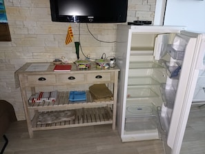 A room with a television mounted on a brick wall, a wooden cabinet with drawers, and a refrigerator with its door open. - Studio 4 persons  (Leucate)