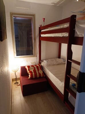 A bunk bed with a red frame, a small bedside table with a lamp, and a window with curtains. - Studio 4 persons  (Leucate)